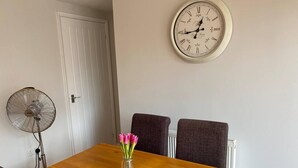 A dining area with a wooden table, two chairs, a vase of pink flowers, a wall clock, and a standing fan.
