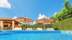 A swimming pool with lounge chairs and umbrellas, surrounded by greenery and a building with a tiled roof.