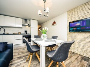A modern kitchen with white cabinets, a dining table with grey chairs, and a TV mounted on a brick wall.
