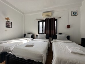 Habitación de hotel con tres camas individuales, una ventana con cortinas, un equipo de aire acondicionado y un ventilador pequeño de pared.