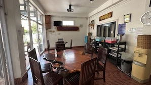 A dining area with a wooden table and chairs, a ceiling fan, and a kitchen area with cabinets and a refrigerator. - VangVieng Santara Hotel (Vang Vieng)