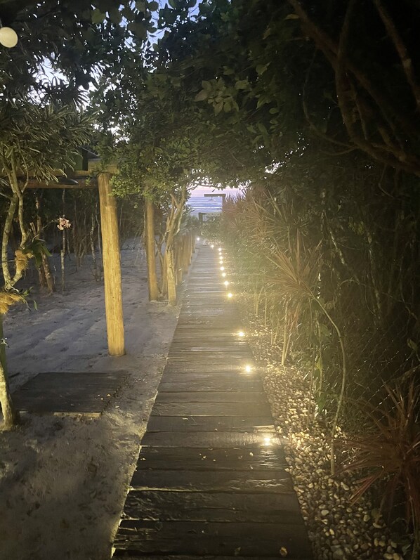 A wooden pathway lined with trees and illuminated by lights, leading to a structure in the distance.