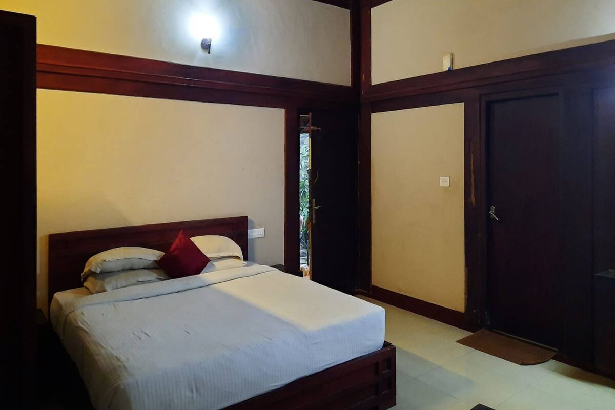 A bedroom with a wooden bed frame, white bedding, and a red pillow. There is a door to the right and a window above the bed.