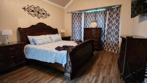 Ein Schlafzimmer mit einem hölzernen Bett, einem Deckenventilator, einem Schminktisch, einem Nachttisch und Vorhängen mit Muster.