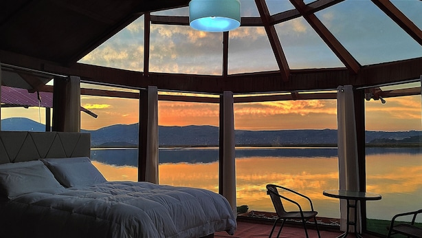 A bedroom with a large bed, a chair, and a table by a window overlooking a lake with mountains in the distance. - Titicaca Los Uros vanalux (Puno)