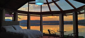 Un dormitorio con una cama grande, una silla y una mesa junto a una ventana con vistas a un lago y montañas al fondo.