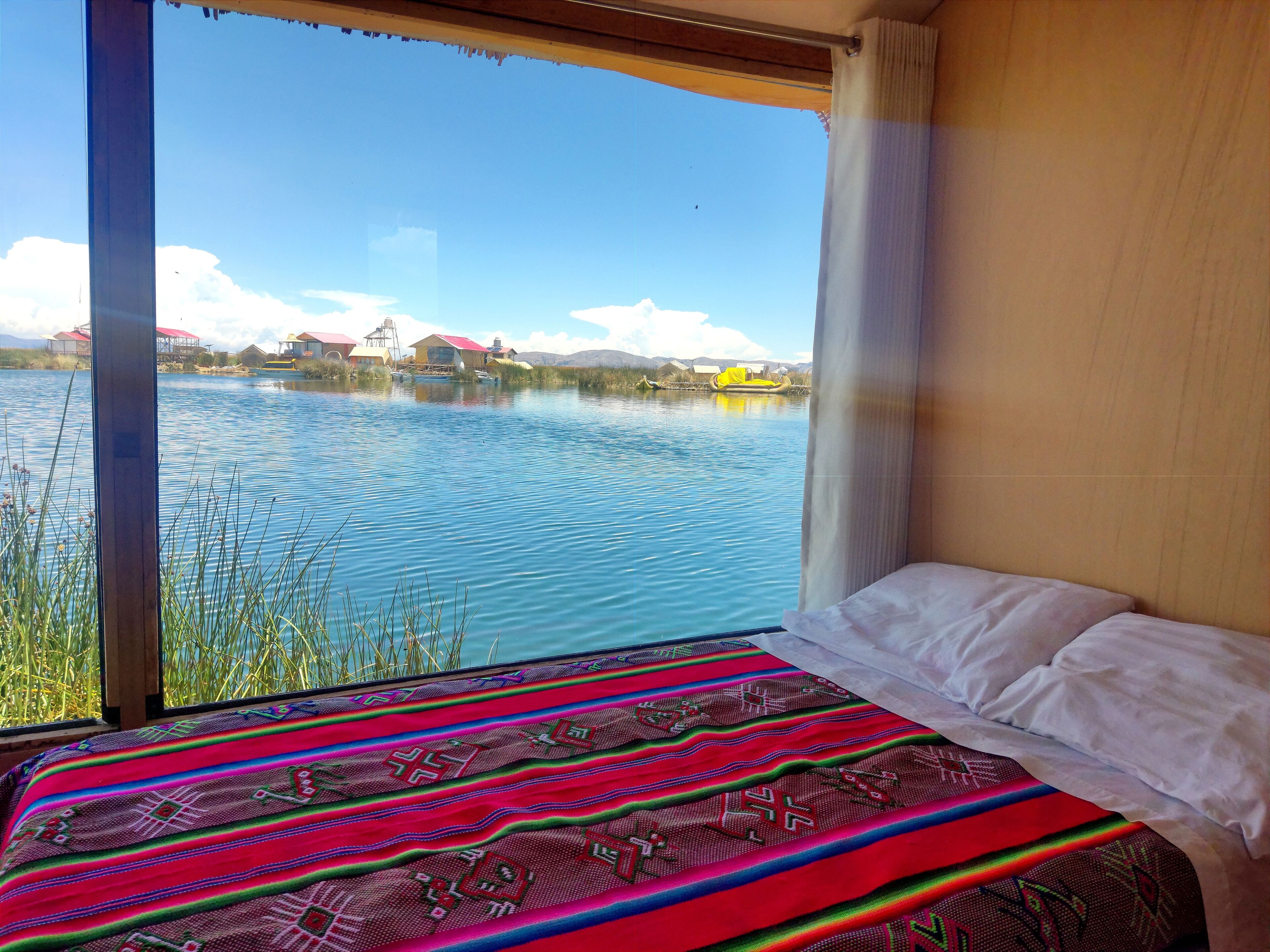 Habitación de hotel con un ventanal grande que da a un lago tranquilo, una cama con manta de colores y vistas a casas y un bote amarillo sobre el agua.