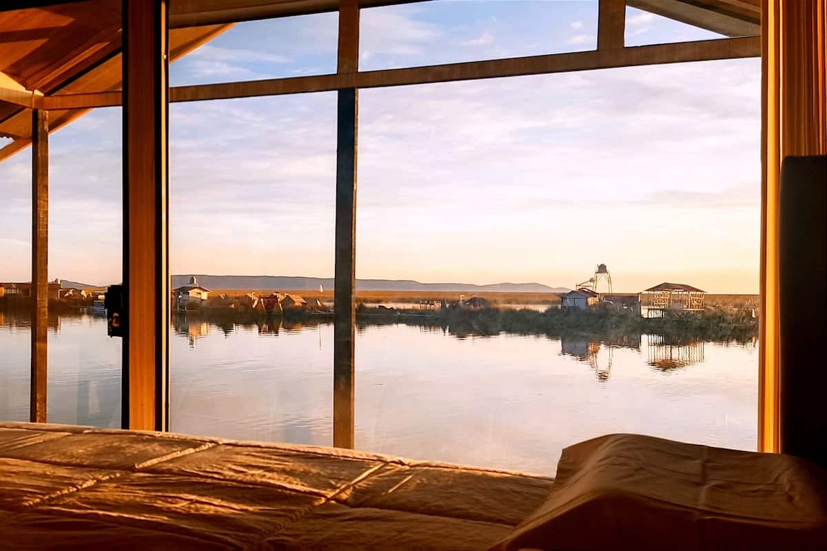 Une pièce dotée d’une grande fenêtre donnant sur un lac paisible, d’un lit et d’une structure de plafond en bois.