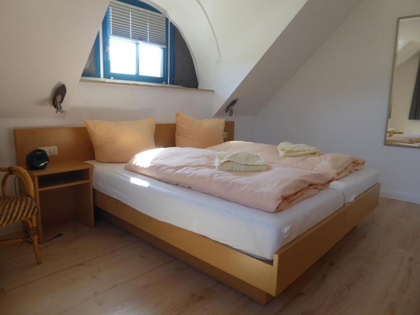 A bedroom with a sloped ceiling, a bed with white bedding, two bedside tables, a chair, and a window with blinds.