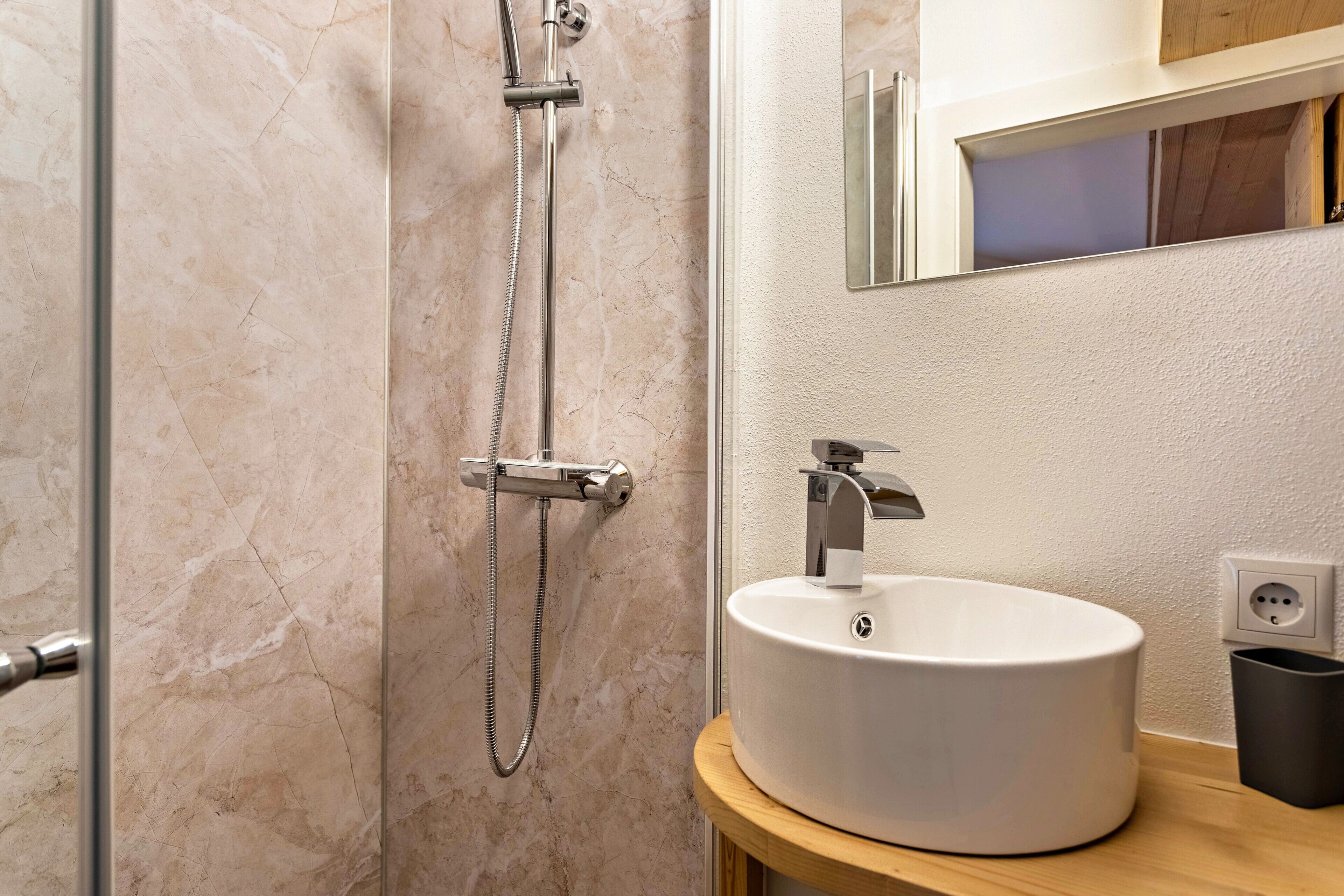 Un baño con pared de mármol, un lavabo blanco y una ducha con cabezal de ducha manual.