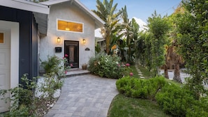 Ein modernes Haus mit einem gepflasterten Auffahrt, die zu einer schwarzen Tür führt, umgeben von üppiger Vegetation und blühenden Pflanzen.