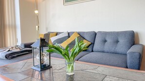 Una sala moderna con un sofá gris, cojines estampados, un florero de vidrio con plantas verdes, un farol negro y un par de gafas sobre una manta doblada.