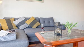 A modern living room with a grey sofa, patterned cushions, a glass coffee table, a small lantern, and a potted plant.