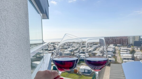 Deux verres à vin remplis de vin rouge sont posés sur un balcon d’où l’on aperçoit un parking et des immeubles.