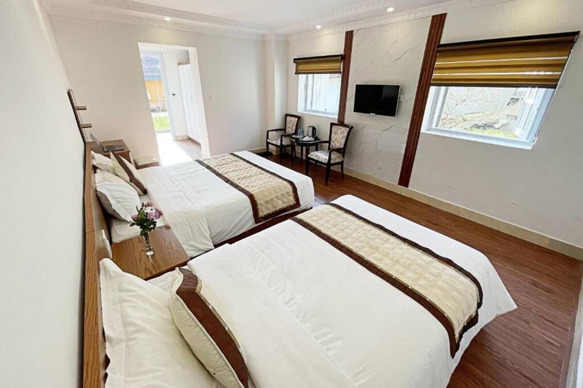 A hotel room with two beds, a TV, a small table with chairs, and a window with blinds.