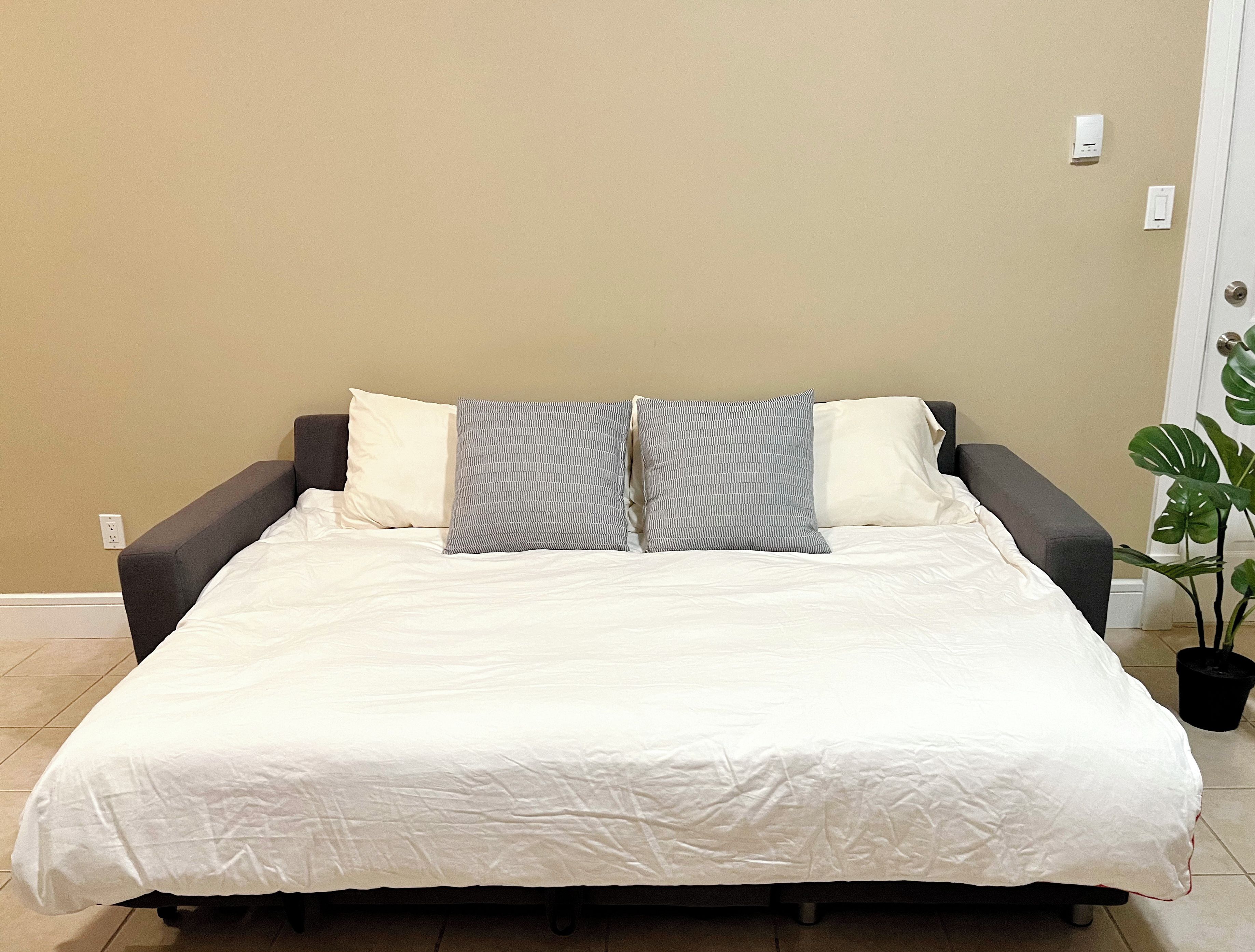 Una cama con ropa de cama blanca, dos almohadas grises y una almohada a rayas grises y blancas.
