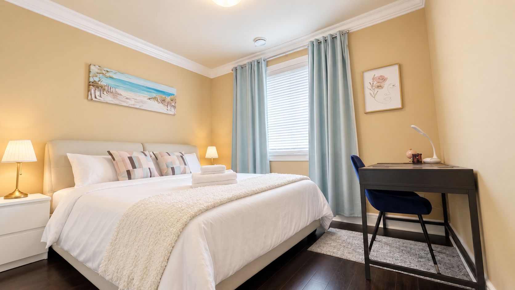 A hotel room with a bed, bedside lamps, a desk with a chair, and artwork on the walls.