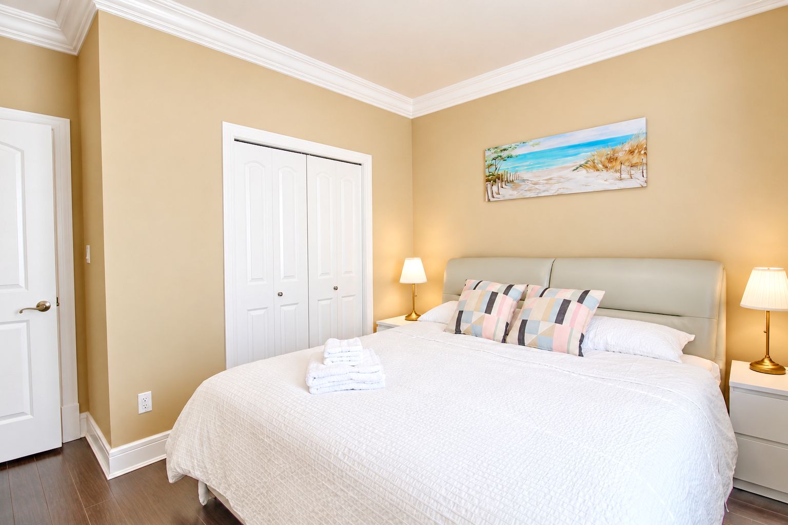 A hotel room with a large bed, two bedside tables with lamps, a painting of a beach scene, and a white door.