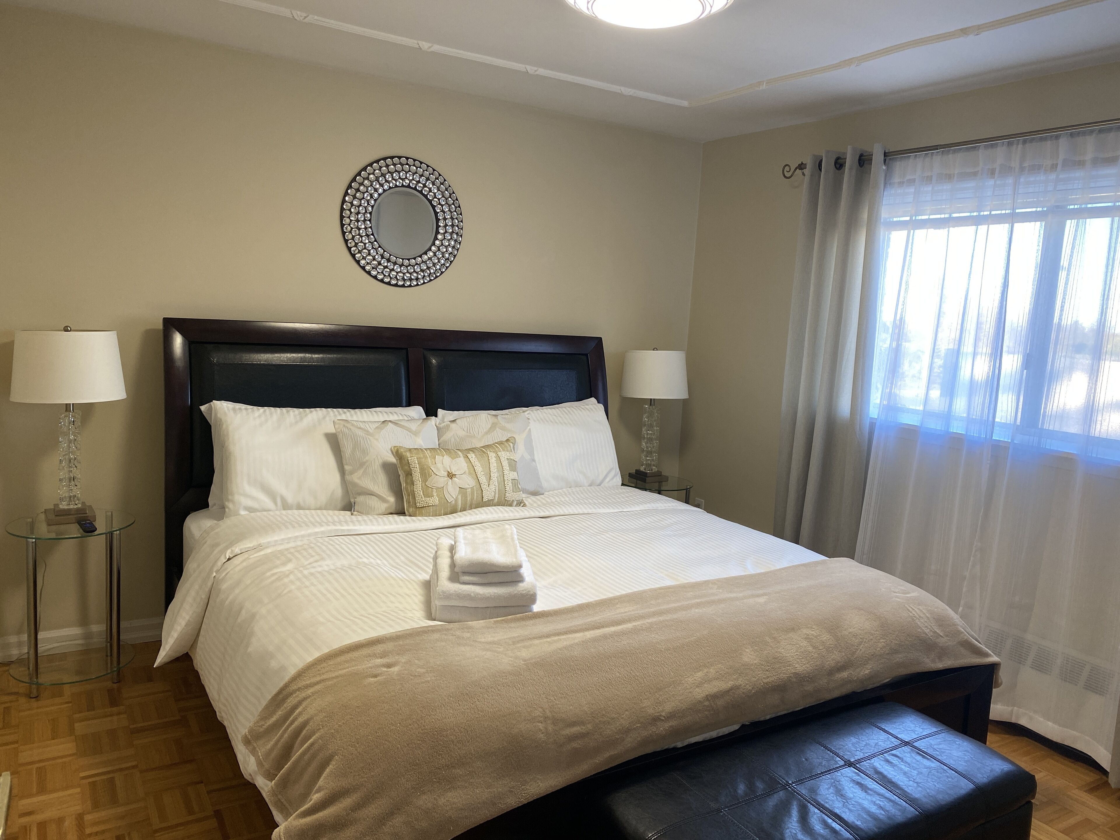 A neatly made bed with a headboard, two bedside lamps, a round mirror, and a window with curtains.