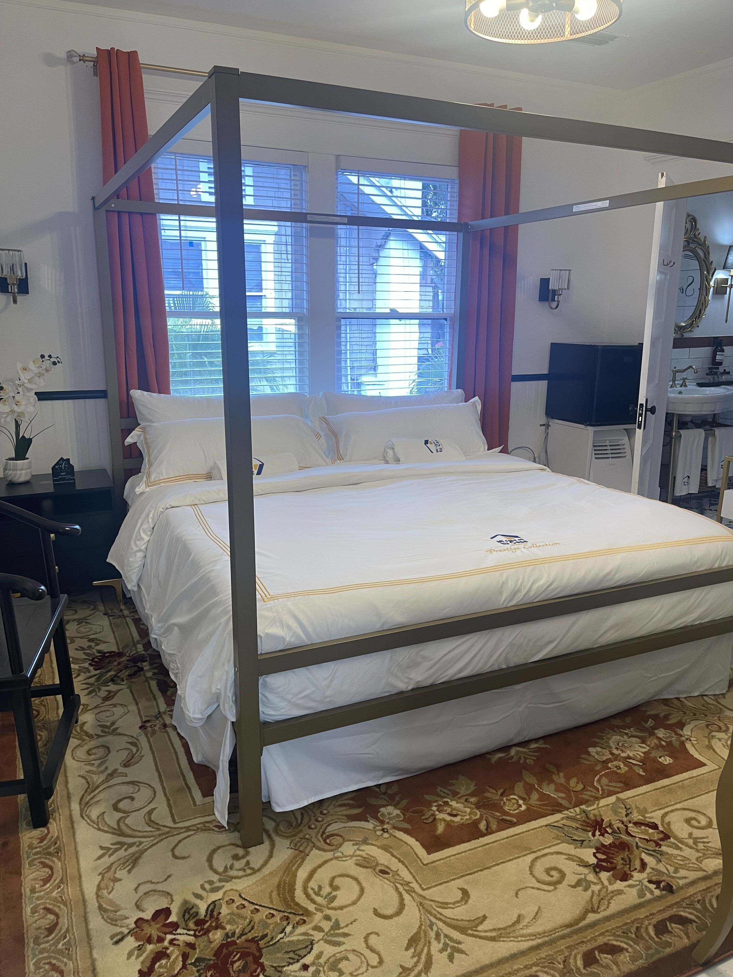 A four-poster bed with white bedding, a floral-patterned rug, and a view of buildings through large windows with red curtains.