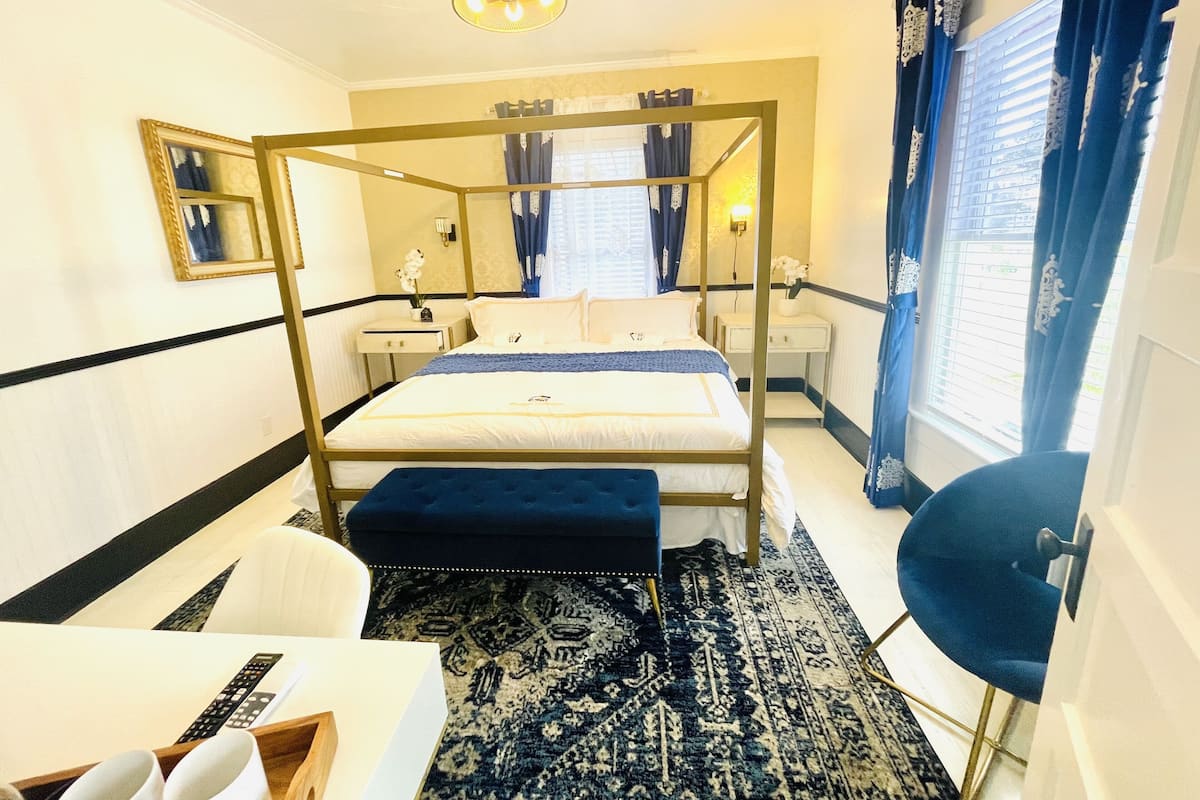 Una habitación de hotel con una cama con dosel, mesitas de noche, un escritorio, una silla, un ventanal grande con cortinas y una alfombra estampada.