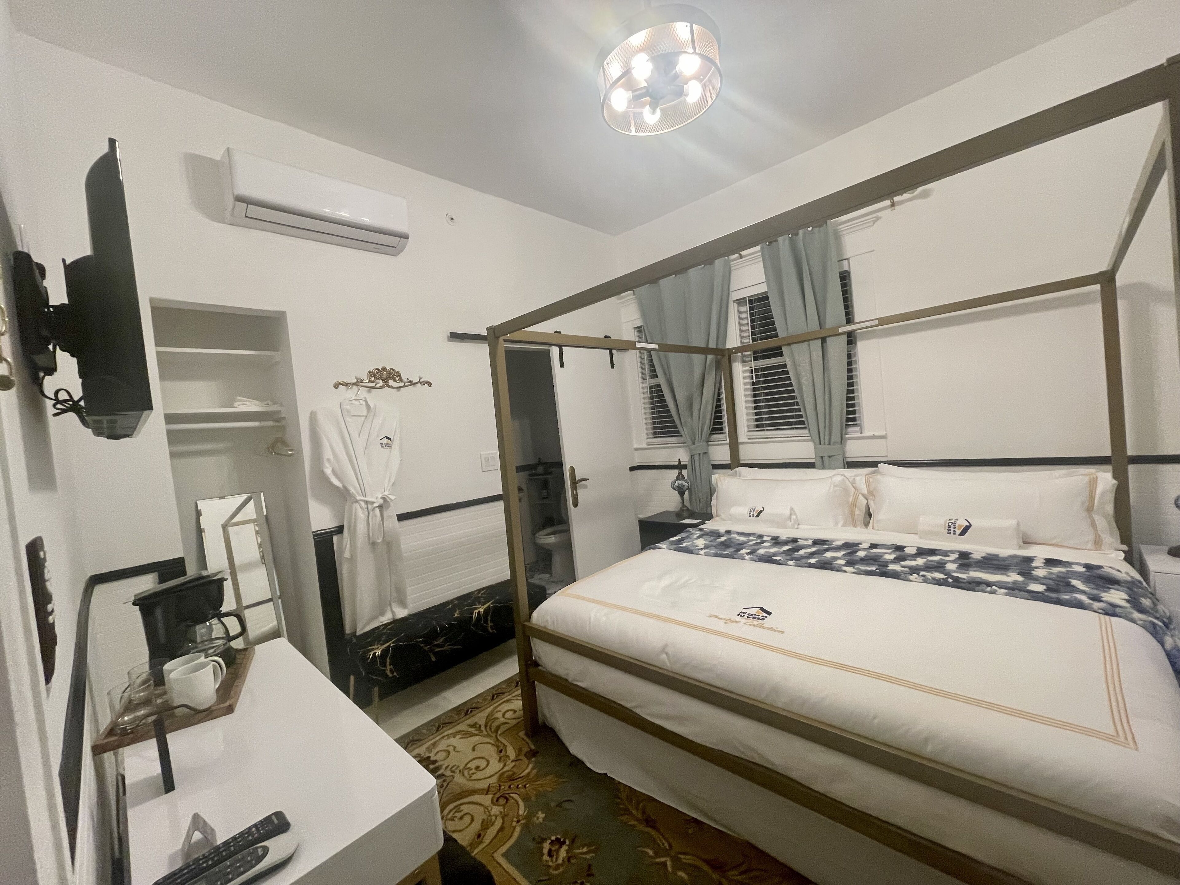 A hotel room with a four-poster bed, a bedside table with a lamp, a robe hanging on the wall, and a small seating area with a table and chairs.