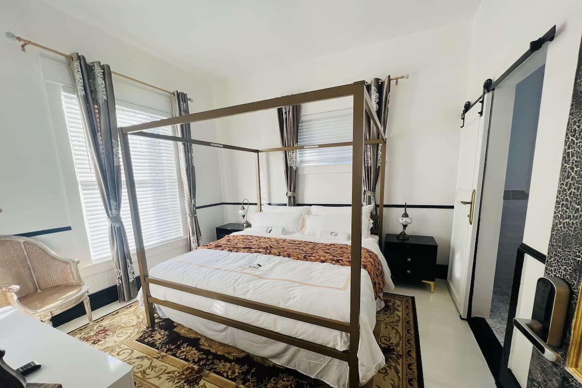 Una cama con dosel, con un edredón blanco y marrón, una mesita de noche con una lámpara y una silla con un cojín estampado.