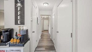 A hotel hallway with a coffee station on the left, featuring a coffee machine, cups, and a coffee bag. The hallway has white walls and doors, leading to a room at the end. - Two bedroom condo for rent! (Columbus)