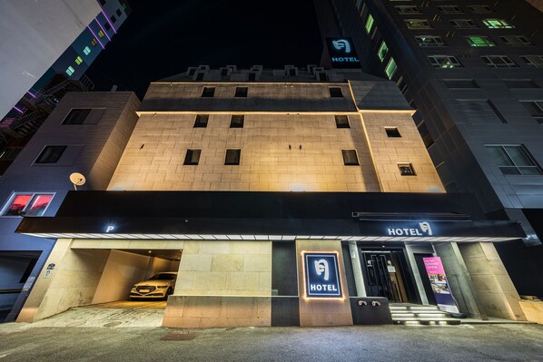 Entrada de un hotel con un letrero, un coche estacionado en un área de estacionamiento techada y una escalera que conduce a la entrada.