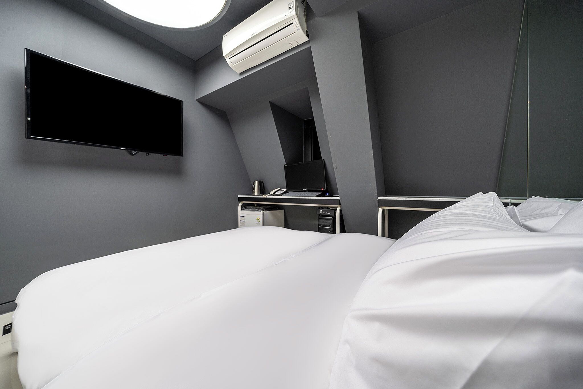 A modern hotel room with a large bed, a flat-screen TV mounted on the wall, a desk with a laptop, and a bedside table with a lamp.