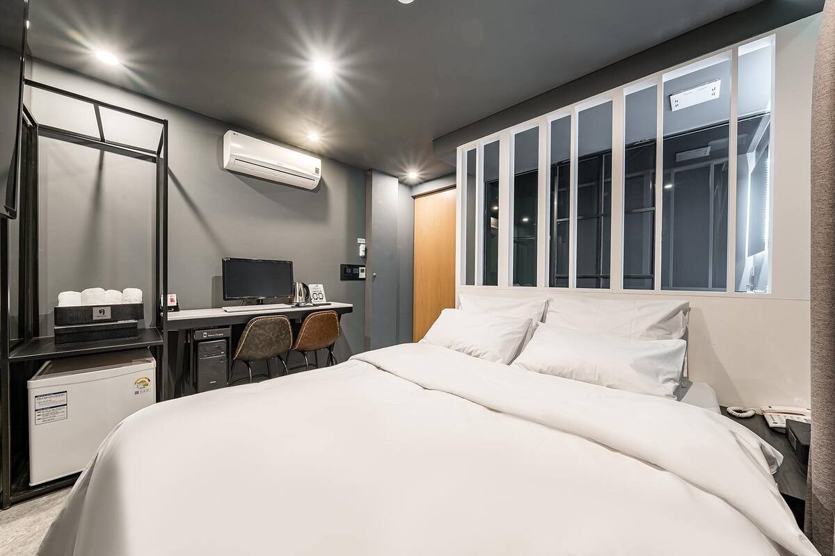 Hotellihuone, jossa on sänky, työpöytä tietokoneella, tuoli, ilmastointilaite ja vaatekaappi.