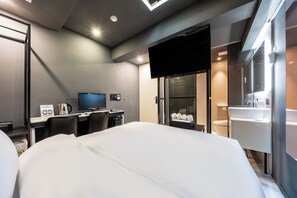 Una habitación de hotel moderna con una cama grande, un escritorio con computadora, un televisor de pantalla plana, un minibar y un baño con espejo y lavamanos.