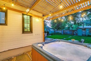A hot tub under a wooden pergola with string lights, a grassy backyard, and a house with a window and a door.