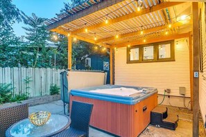 A hot tub under a wooden pergola with string lights, surrounded by a wooden fence and a house.