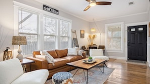 A living room with a brown leather sofa, a white armchair, a wooden coffee table, and a patterned ottoman. There is a ceiling fan, a black door, and a window with blinds. - 4 min walk to Town Square! 3bd 2bath cozy Historical home (Georgetown)
