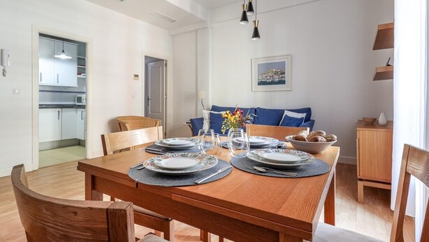 Un coin repas avec une table en bois dressée pour quatre personnes, un canapé avec des coussins bleus et une cuisine visible au fond.