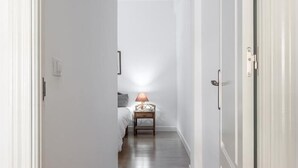 Un couloir étroit, avec un lit et une table de chevet au fond, agrémenté d’une lampe et d’un interrupteur fixé au mur.
