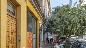 Une rue étroite bordée de scooters et de voitures garées, un immeuble jaune avec une grande porte en bois et un trottoir bordé d’arbres.
