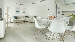 Un salon moderne et bien éclairé, avec un coin repas, des meubles blancs et une cuisine en arrière-plan.