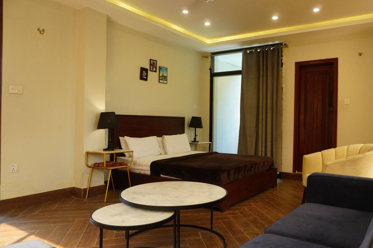 Habitación de hotel con cama, dos mesitas de noche con lámparas, un sofá, una pequeña mesa redonda y una ventana con cortinas.