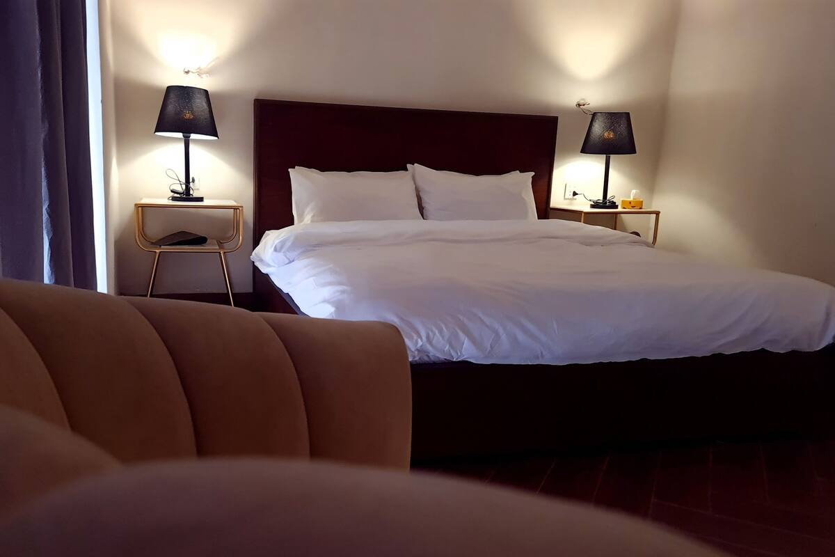 Habitación de hotel con una cama grande, dos mesitas de noche con lámparas, una silla pequeña y vistas al exterior a través de una ventana.