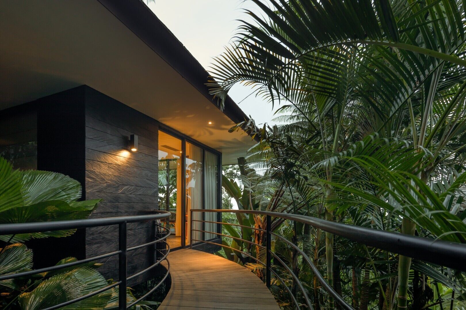 A modern building with a stone facade, a glass entrance, and a wooden walkway surrounded by lush tropical vegetation.