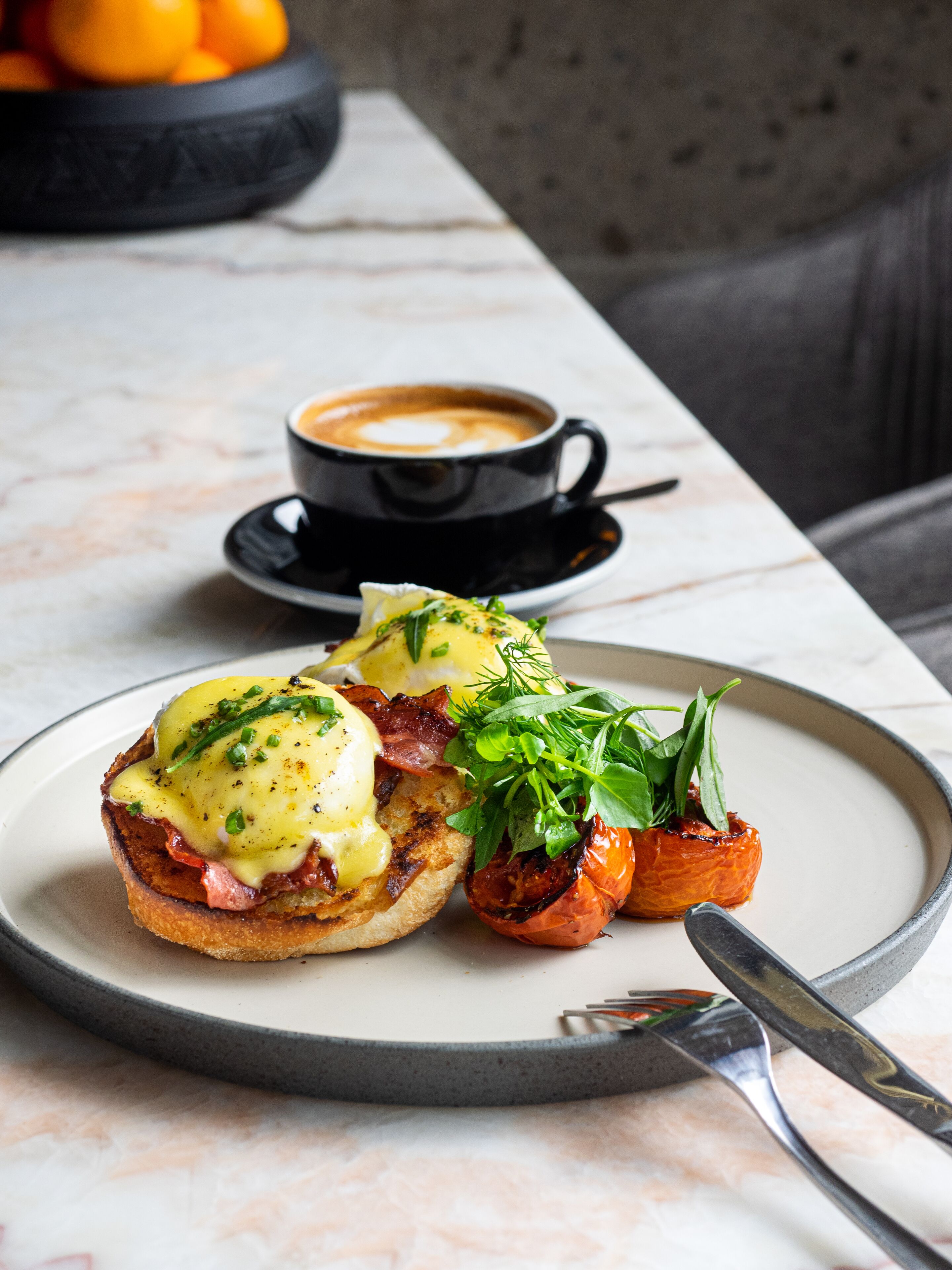 Un plato de desayuno con huevos benedictinos, tomate asado al lado y una taza de café sobre una mesa de mármol.