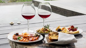 Una mesa con diversas comidas y dos copas de vino tinto, sobre una terraza de madera con vistas panorámicas a un área boscosa.