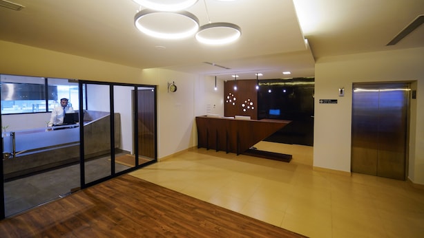 Una zona de recepción moderna con suelo de madera, un mostrador de recepción y una mesa de recepción. Hay un ascensor a la derecha y una escalera que sube a otro piso.
