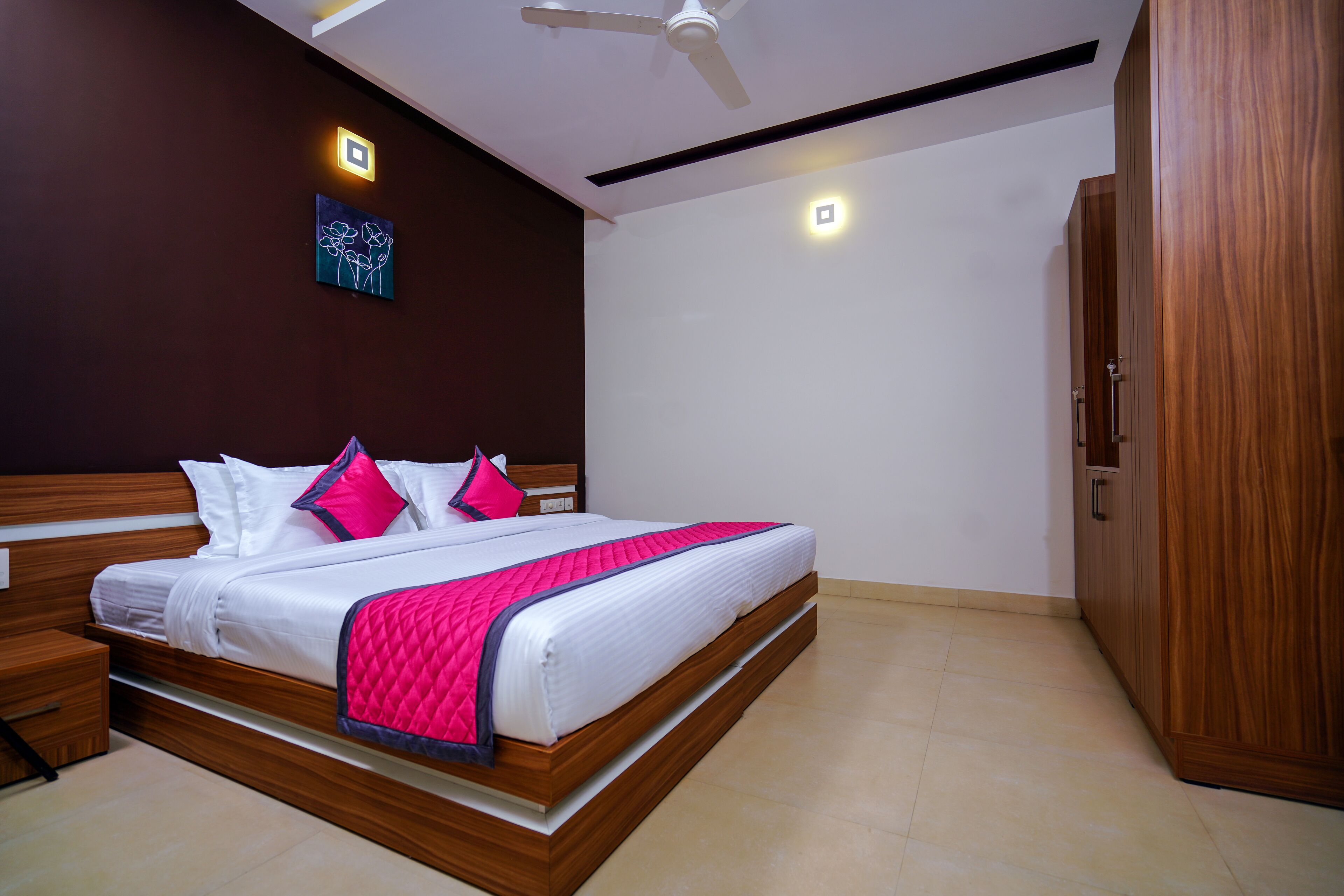 Cama bem arrumada com roupa de cama branca e rosa, cabeceira de madeira e mesa de cabeceira com gaveta.