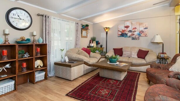 Un salon chaleureux avec un canapé beige, une table basse et un tapis rouge. Il y a une horloge au mur et une plante sur la table d’appoint.