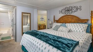 Une chambre à coucher avec un lit, une tête de lit en bois, un élément décoratif fixé au mur, et une salle de bain visible à travers une porte ouverte.