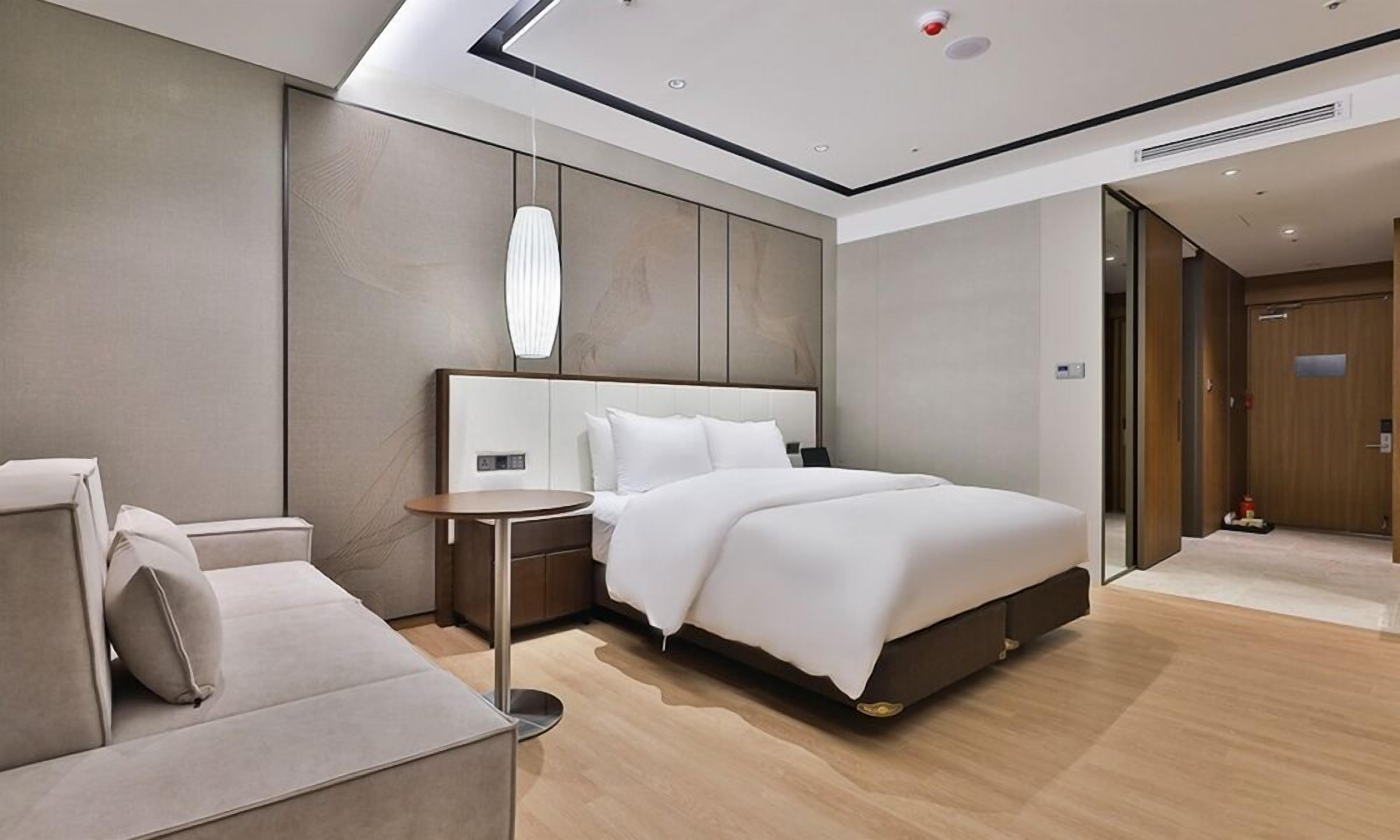 A modern hotel room with a large bed, a bedside table, a small round table, and a sofa.