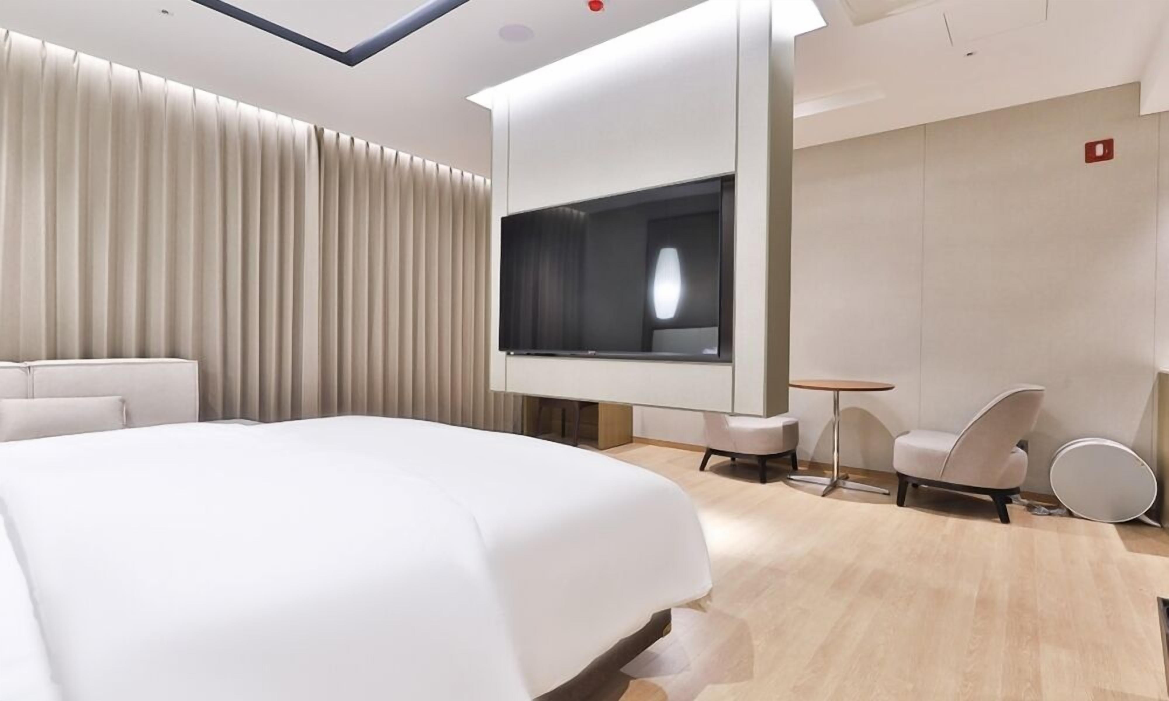A modern hotel room with a large bed, a TV mounted on the wall, a small round table, and two chairs.